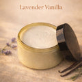 Open jar of lavender vanilla product with a black lid, surrounded by lavender flowers and vanilla beans on a beige background.